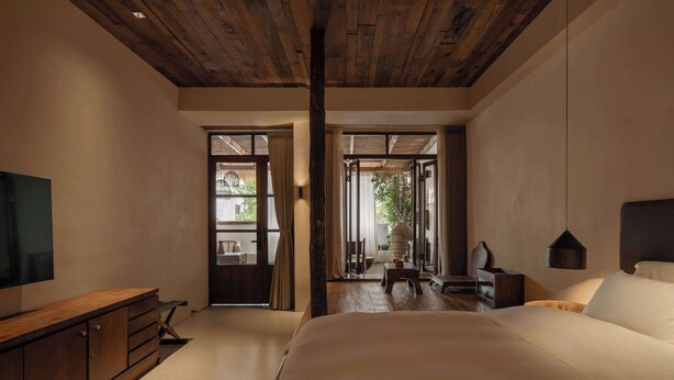 A bedroom with a wooden ceiling, a large bed, a TV on the left, and a view of the outdoors through a glass door. - YueLore Retreat (Chengdu)