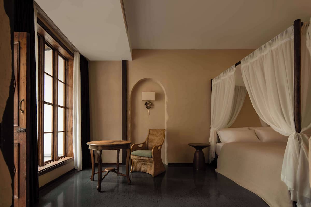 A bedroom with a large window, a chair, a bedside table, and a canopy bed.
