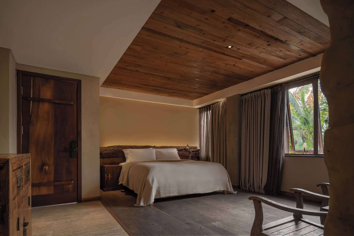 A bedroom with a wooden ceiling, a bed with white linens, a nightstand, a wooden chair, and a window with curtains.