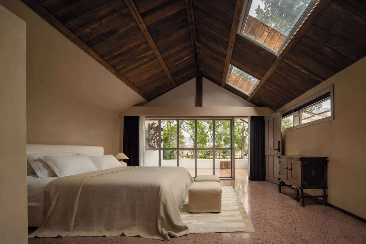 A bedroom with a large bed, a bench, a wooden cabinet, and a view of greenery through large windows.