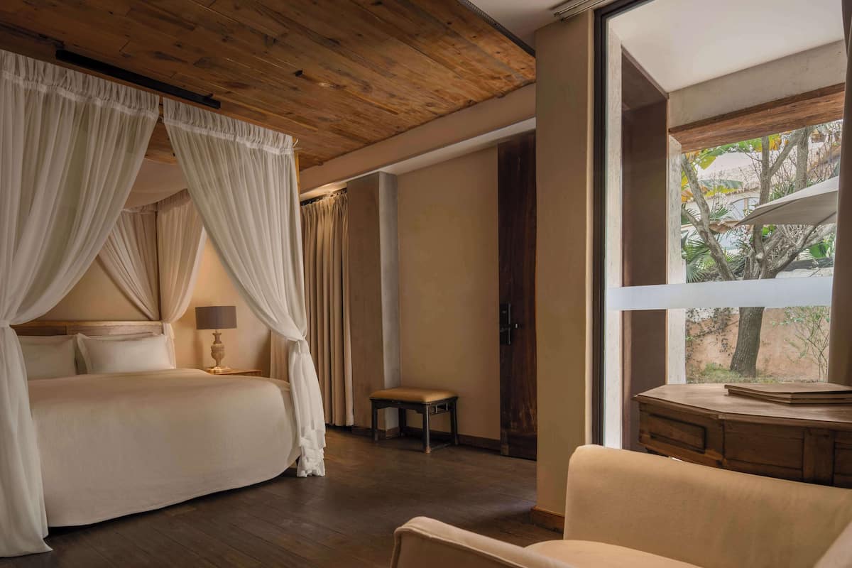 A bedroom with a canopy bed, a wooden ceiling, a large window, and a desk with a lamp.