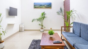 Una sala de estar con sofá, una mesa de centro, un televisor montado en la pared y varias plantas en macetas.