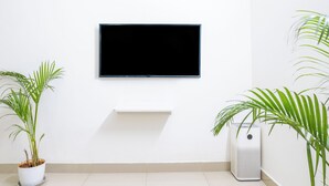 Una habitación con un televisor fijado en la pared, un estante blanco debajo y dos macetas a cada lado.