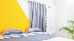 Una cama con colcha blanca y azul, dos almohadas y una cortina gris.