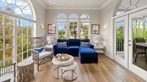 A spacious living room with a blue sectional sofa, a round coffee table, and a wicker chair. Large windows offer views of the outdoors. - Gorgeous New pool home a bike ride from beach! (Sanibel)