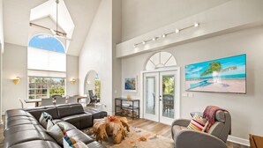 A spacious living area with a large sectional sofa, a coffee table, and a gray armchair. There is a dining area with a table and chairs, and a large window providing natural light. - Gorgeous New pool home a bike ride from beach! (Sanibel)