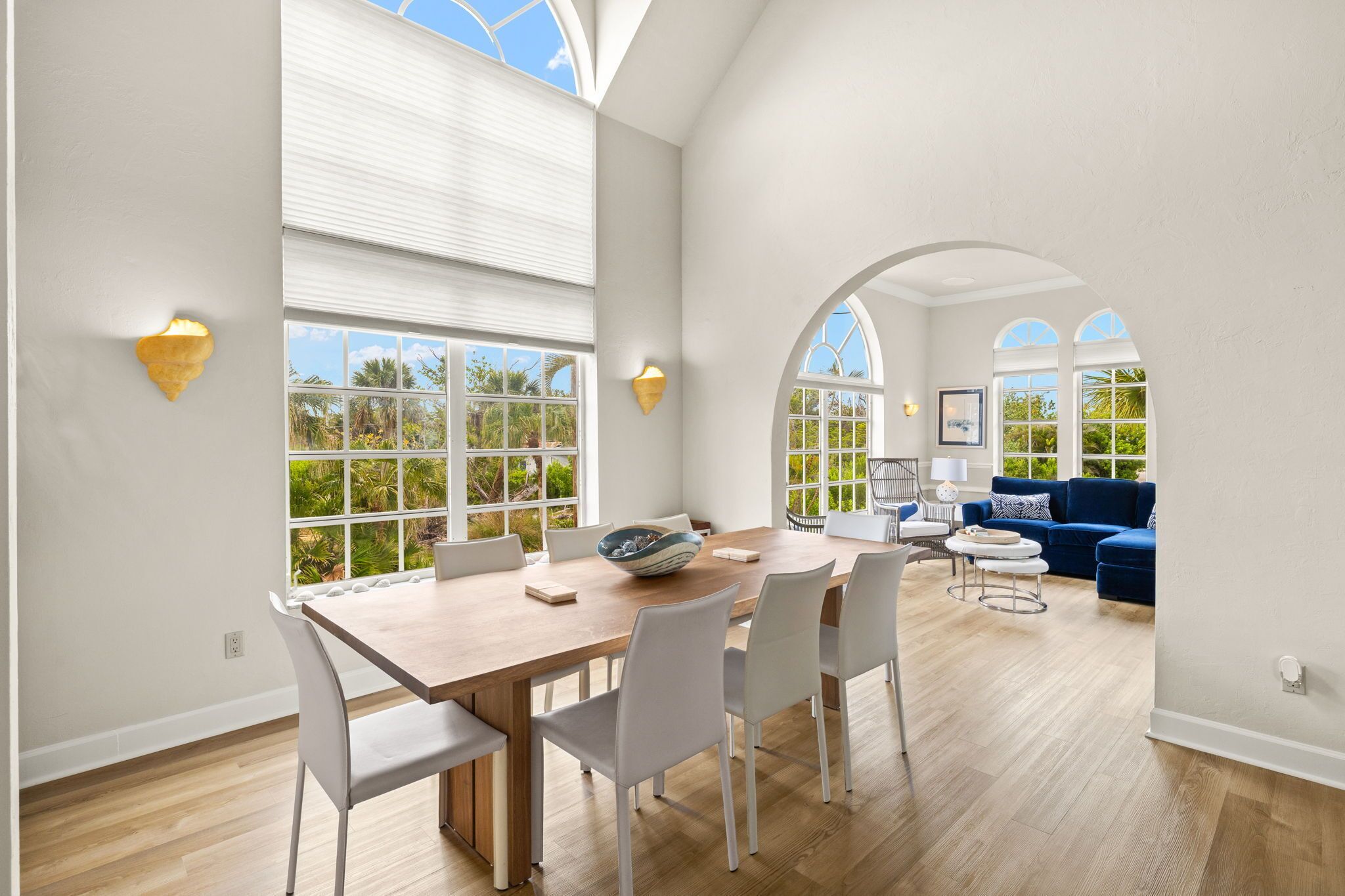 A spacious dining area with a wooden table and white chairs, large windows with views of greenery, and a blue sofa in the background.