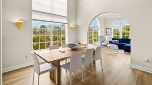 A spacious dining area with a wooden table and white chairs, large windows with views of greenery, and a blue sofa in the background. - Gorgeous New pool home a bike ride from beach! (Sanibel)