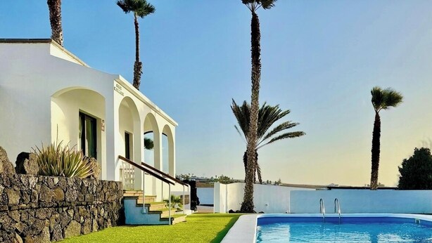 A modern pool area with a stone wall, a white building with arches, and palm trees.