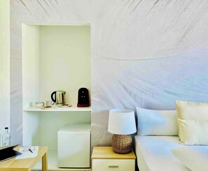 Ein Hotelzimmer mit einem Bett, einem Nachttisch mit Lampe, einem kleinen Kühlschrank, einem Regal mit Wasserkocher und Kaffeemaschine sowie einer Wandablage mit Flasche und Tasse.