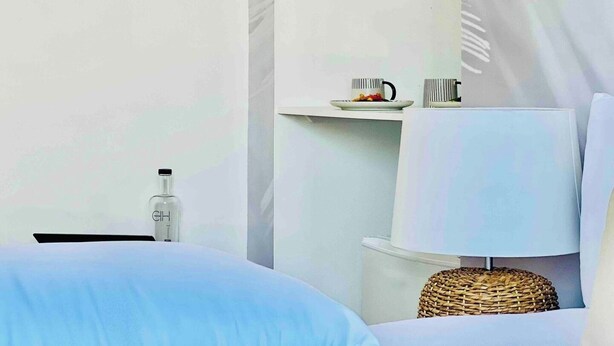 Habitación de hotel minimalista con una cama, una mesita de noche con una botella, una lámpara y un pequeño estante con un plato y dos tazas.
