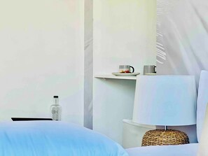 Une chambre d’hôtel minimaliste comprenant un lit, une table de chevet avec une bouteille, une lampe et une petite étagère avec une assiette et deux tasses.