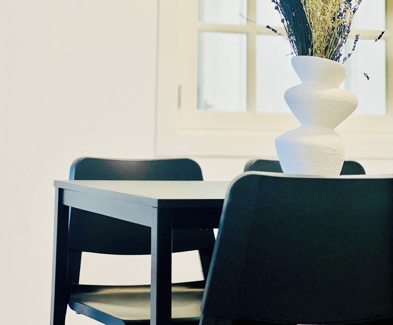 Una mesa de comedor con sillas, un jarrón blanco con flores y una ventana al fondo.