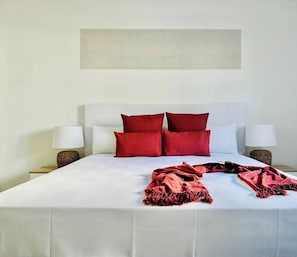 Un lit bien fait, avec du linge de lit blanc et des coussins rouges, deux lampes de chevet et un mur sobre orné d’une œuvre d’art à la texture intéressante, placée au-dessus du lit.