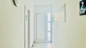 Un couloir avec une porte au fond, une plaque indiquant le numéro de la pièce (2), et un interrupteur sur le mur.