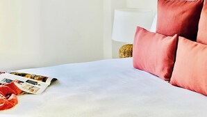 Una cama bien hecha con sábanas blancas y almohadas rojas, una revista sobre la cama y una lámpara al fondo.