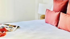 A neatly made bed with white linens and red pillows, a magazine on the bed, and a lamp in the background.