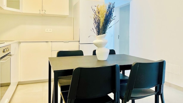 A modern kitchen with white cabinets, a dining table with four chairs, and a vase with dried flowers on the table.