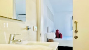 A hotel bathroom with a sink, mirror, and a door leading to a bedroom with a bed, pillows, and a nightstand.