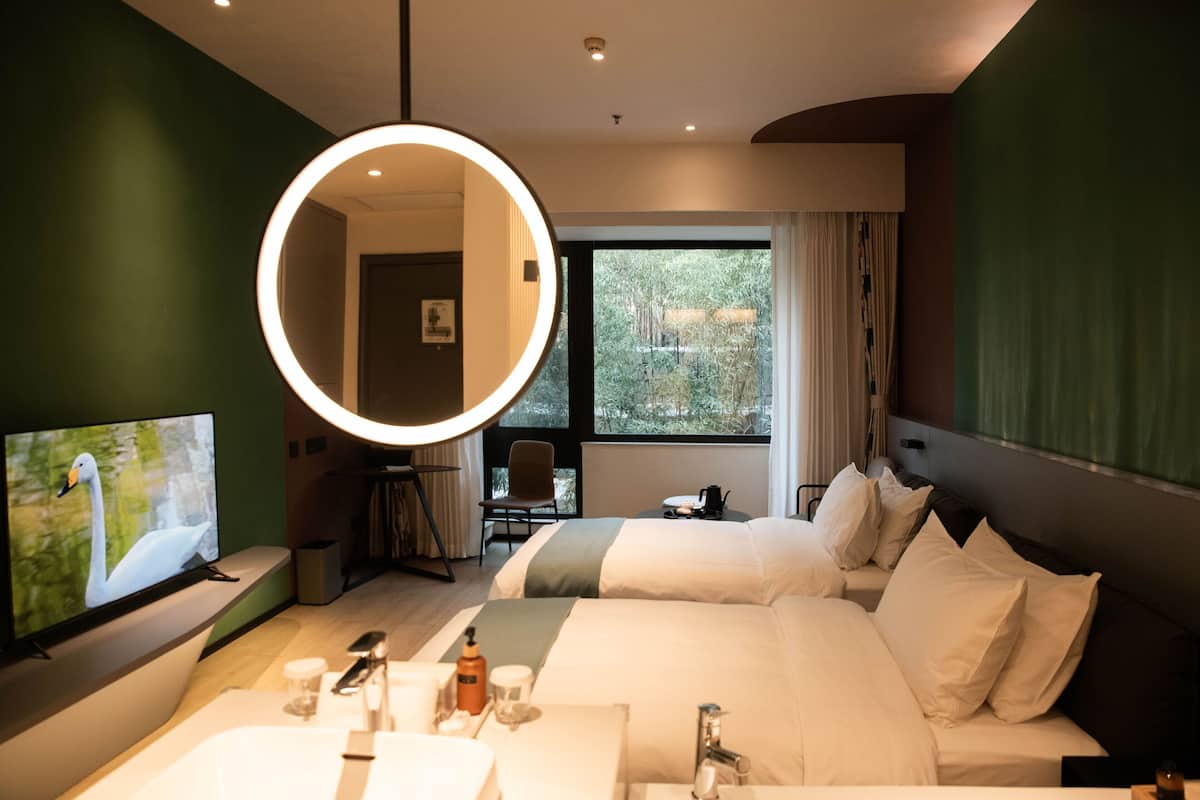 A hotel room with two beds, a TV, a round mirror, a desk, and a window with a view of greenery.