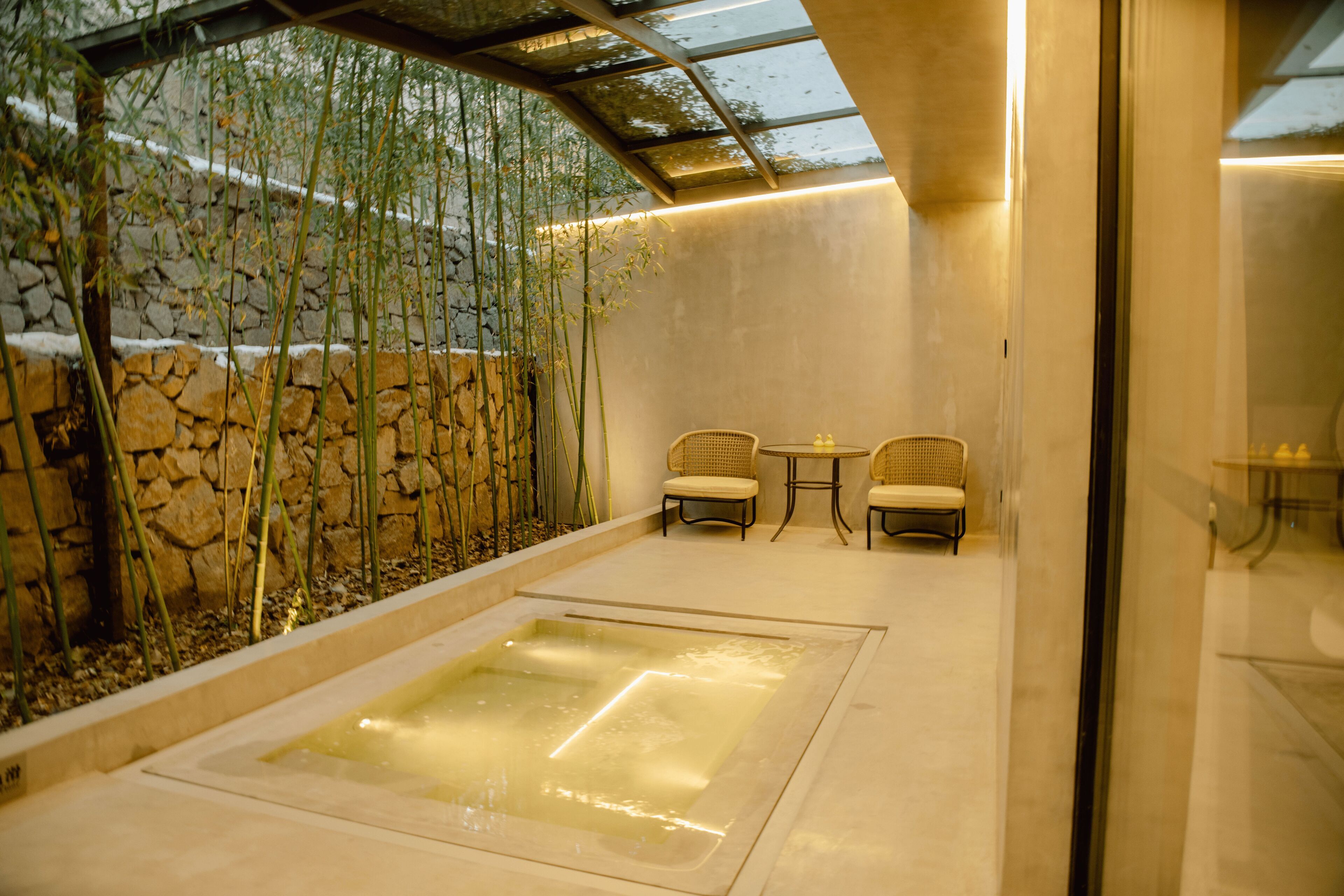 Un espacio de spa con un jacuzzi, pared de piedra, plantas de bambú y dos sillas de mimbre.