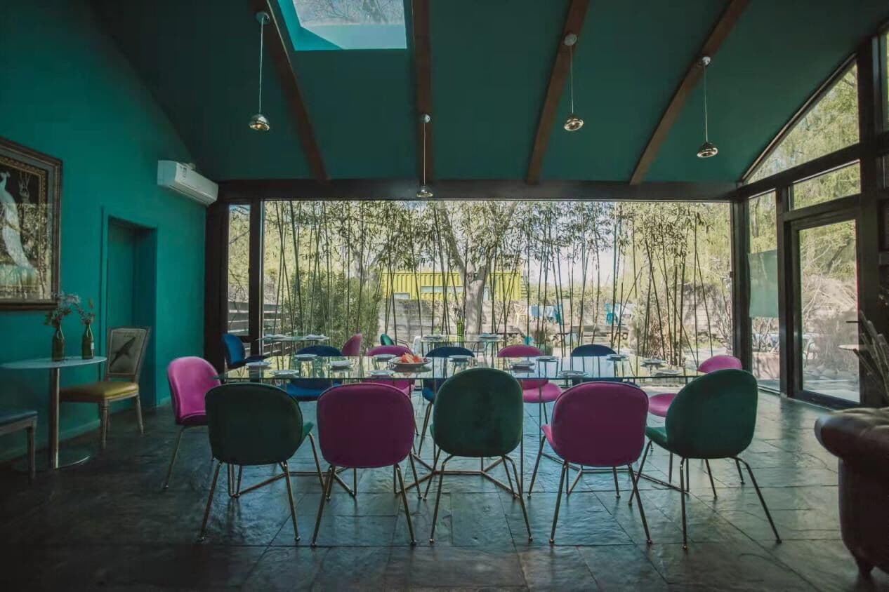 A spacious room with a large dining table surrounded by colorful chairs, large windows offering a view of trees, and a ceiling with wooden beams and hanging lights.