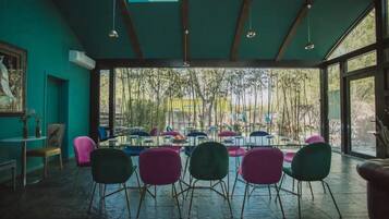 A spacious room with a large dining table surrounded by colorful chairs, large windows offering a view of trees, and a ceiling with wooden beams and hanging lights.