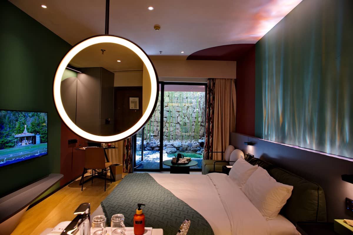 A hotel room with a large bed, a round mirror, a TV, a dining table, and a view of a garden through the glass doors.