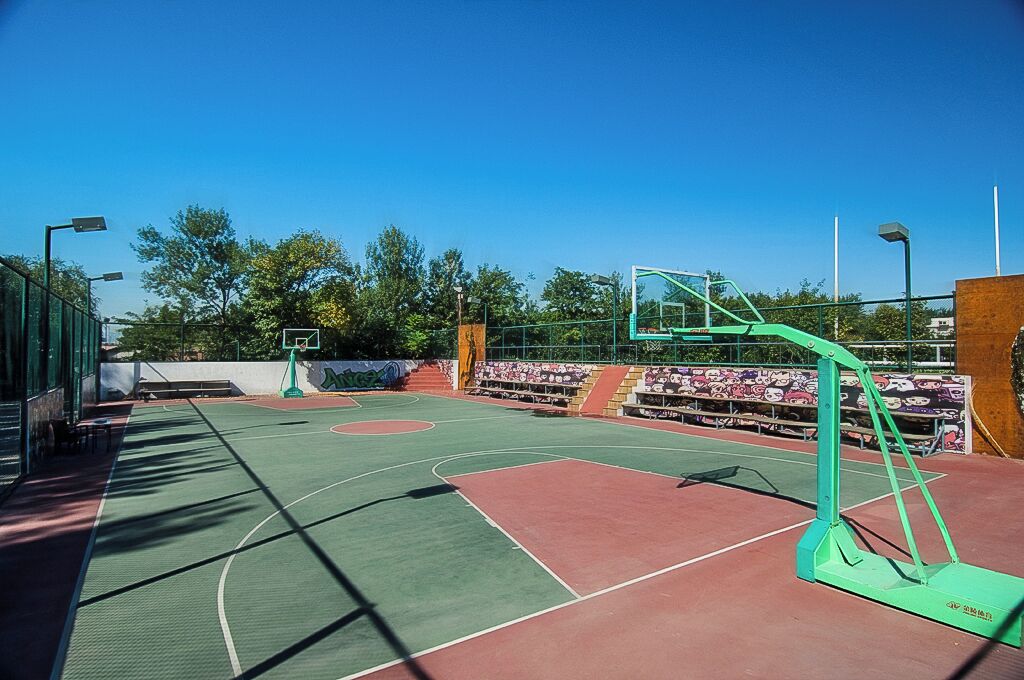 An outdoor basketball court with a green and red playing surface, a basketball hoop, and bleachers in the background.