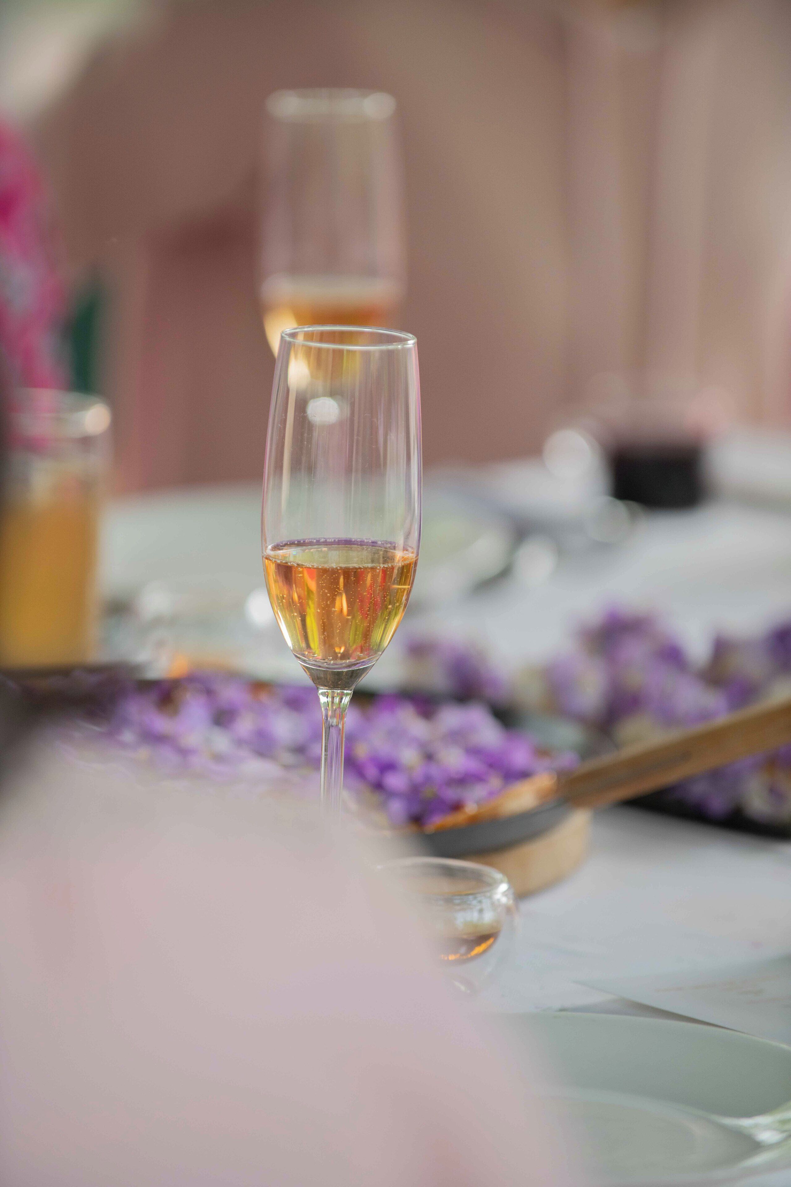 A table with a glass of white wine, a spoon, and scattered purple flowers.