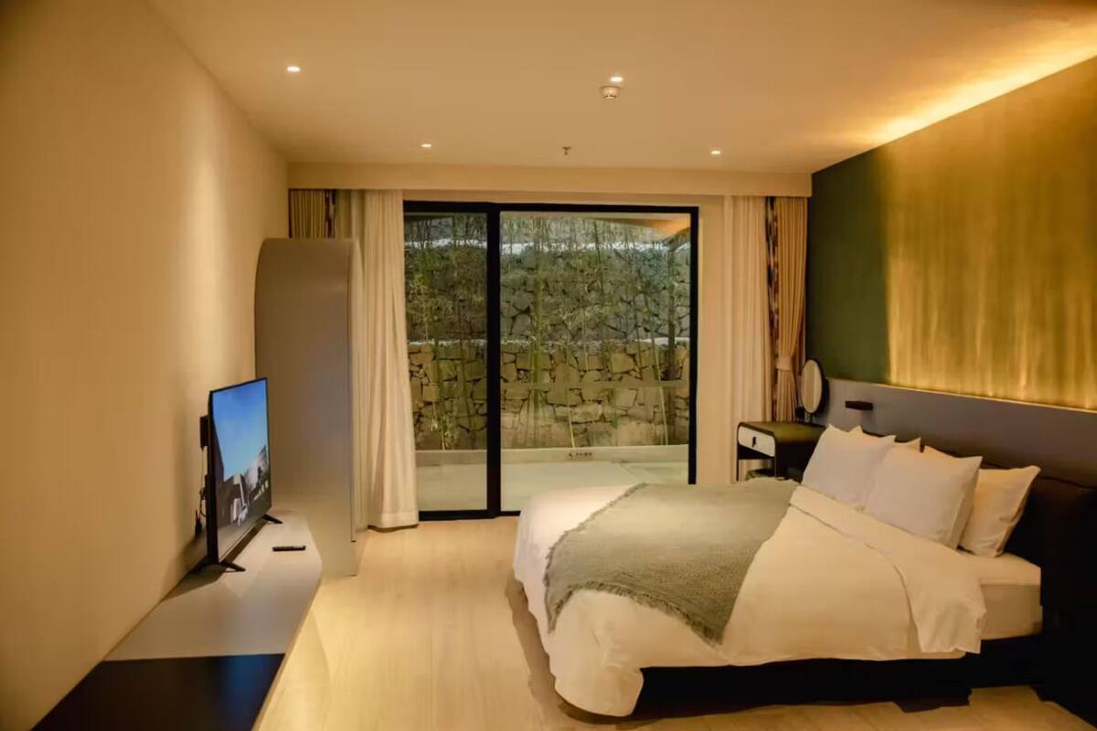 A modern hotel room with a large bed, a flat-screen TV, a nightstand, and a view of a stone wall through a glass door.
