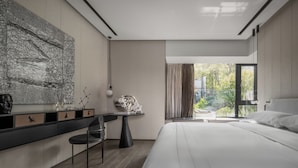 Ein modernes Schlafzimmer mit einem großen Bett, einem Schreibtisch mit Stuhl, einem großen Fenster und Blick auf Grünflächen.