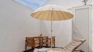 Um deck de madeira com uma poltrona bege e uma mesa com cadeiras. Um guarda-sol está aberto sobre a poltrona.