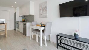 Uma cozinha moderna com armários brancos, uma mesa de jantar com cadeiras, uma TV preta na parede e uma pequena mesa com uma planta em vaso.