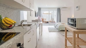 Cozinha moderna com bancada de granito, pia e micro-ondas. Possui área de jantar com mesa e cadeiras, e uma cama com vista para o exterior.