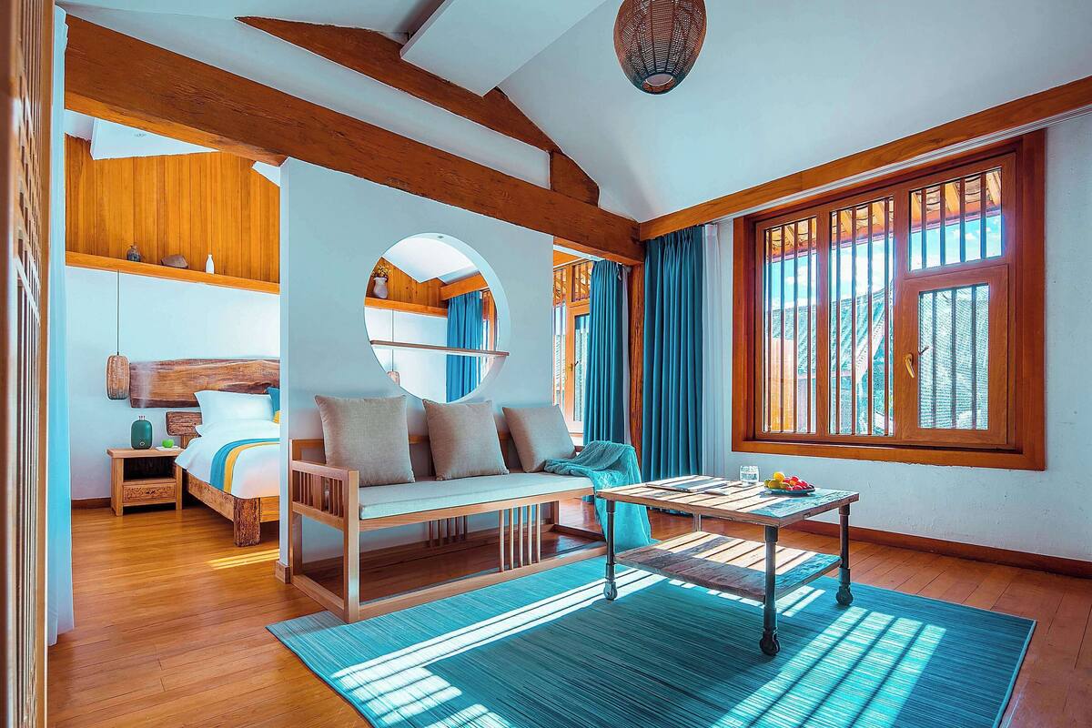 Ein modernes Zimmer mit Holzbalkendecke, einem runden Spiegel, einem Bett mit blau-weiß gestreifter Bettdecke, einer hölzernen Bank, einem kleinen Tisch mit Obstschale und einem großen Fenster mit Vorhängen.