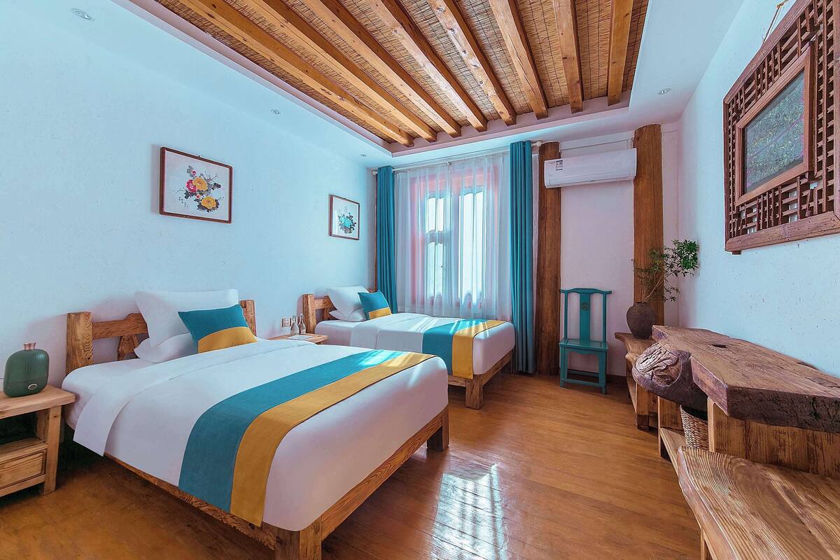 Ein Hotelzimmer mit zwei Betten, Holzboden, Holzbalkendecke und einem Vorhangfenster.