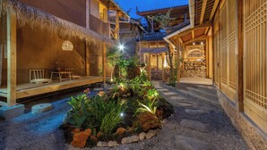 A traditional courtyard with a thatched roof seating area, stone pathway, and a small water feature surrounded by plants.