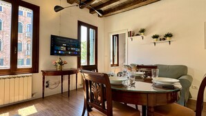 A dining area with a wooden table set for two, a green sofa, and a TV mounted on the wall displaying a menu. - Ca' Angelica Canal View (Venecia)