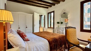 A bedroom with a bed, a chair, a dresser, a mirror, and a window with a view of buildings. - Ca' Angelica Canal View (Venecia)