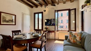 A dining area with a round table set for four, wooden chairs, a sofa with a geometric cushion, and a view of a red brick building through a window. - Ca' Angelica Canal View (Venecia)