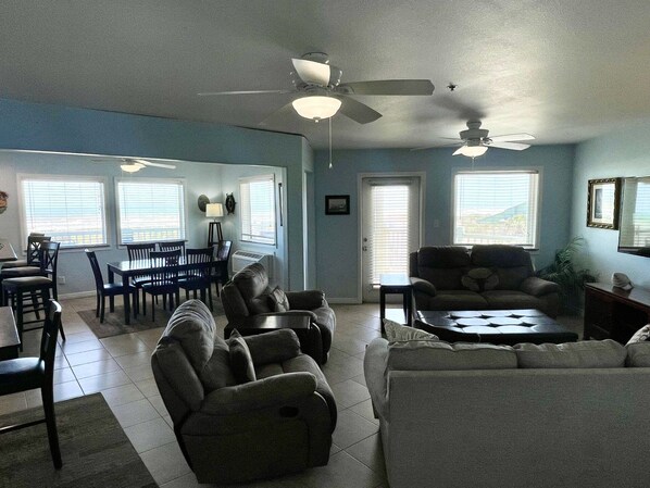 A living room with a dining area, two ceiling fans, and large windows.