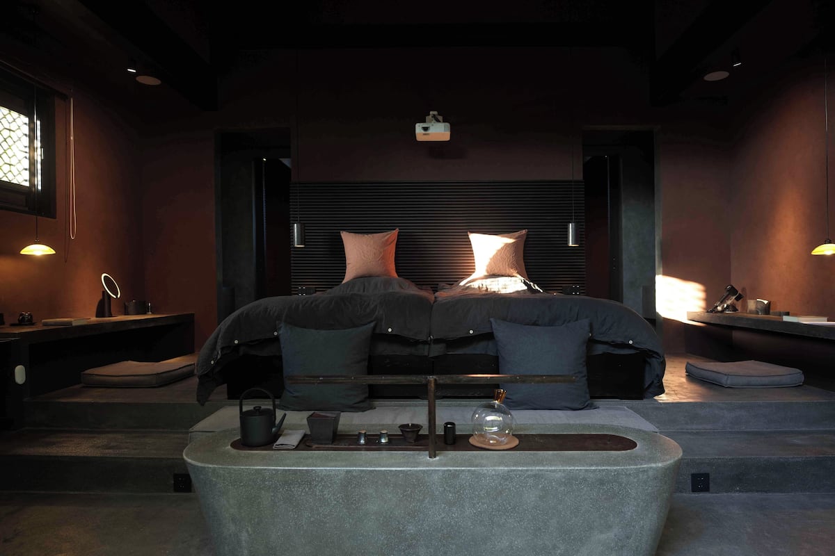 A modern bedroom with a large bed, bedside tables, a reading area, and a stone bathtub.