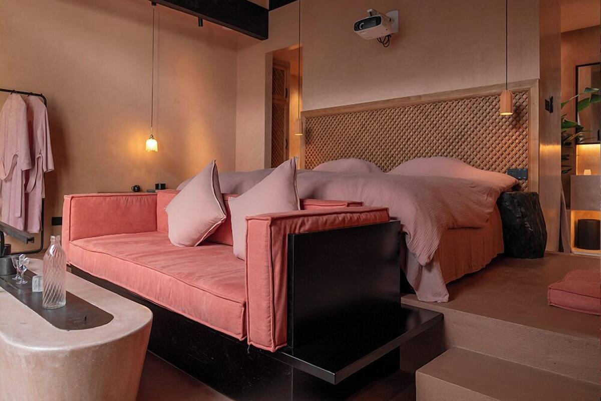 A modern bedroom with a pink sofa, a bed with a wooden headboard, a small table with a vase, and a wall-mounted projector.