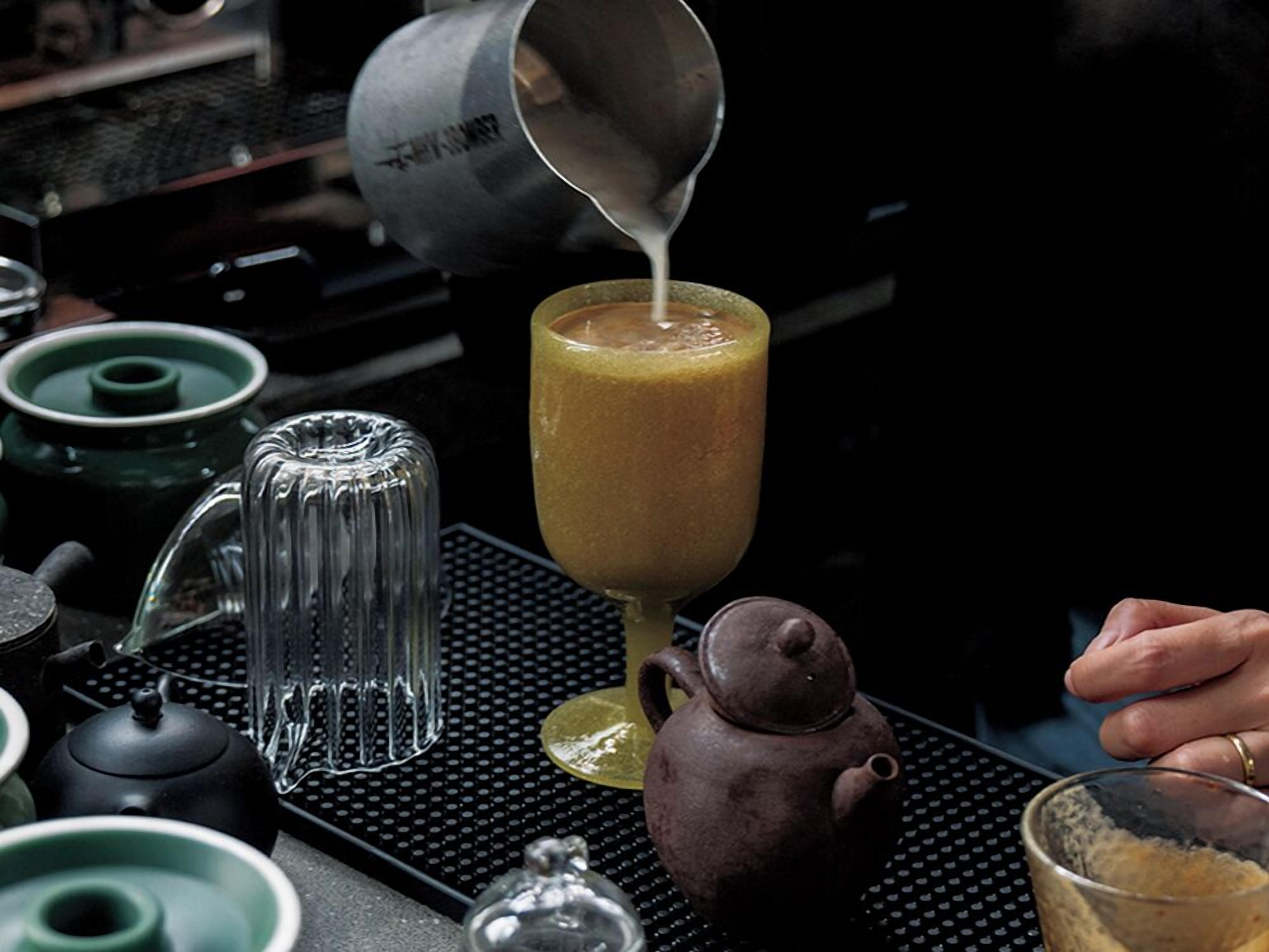 A kitchen with a coffee maker, a teapot, and a glass of a yellowish beverage being poured.