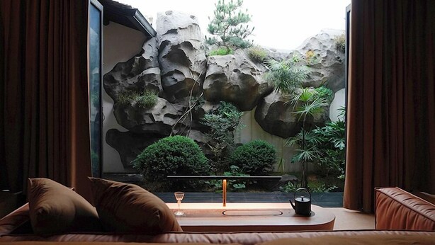 A room with a bed, a bedside table with a glass, and a view of a rock garden with plants. - Stayinmind (Xuancheng)