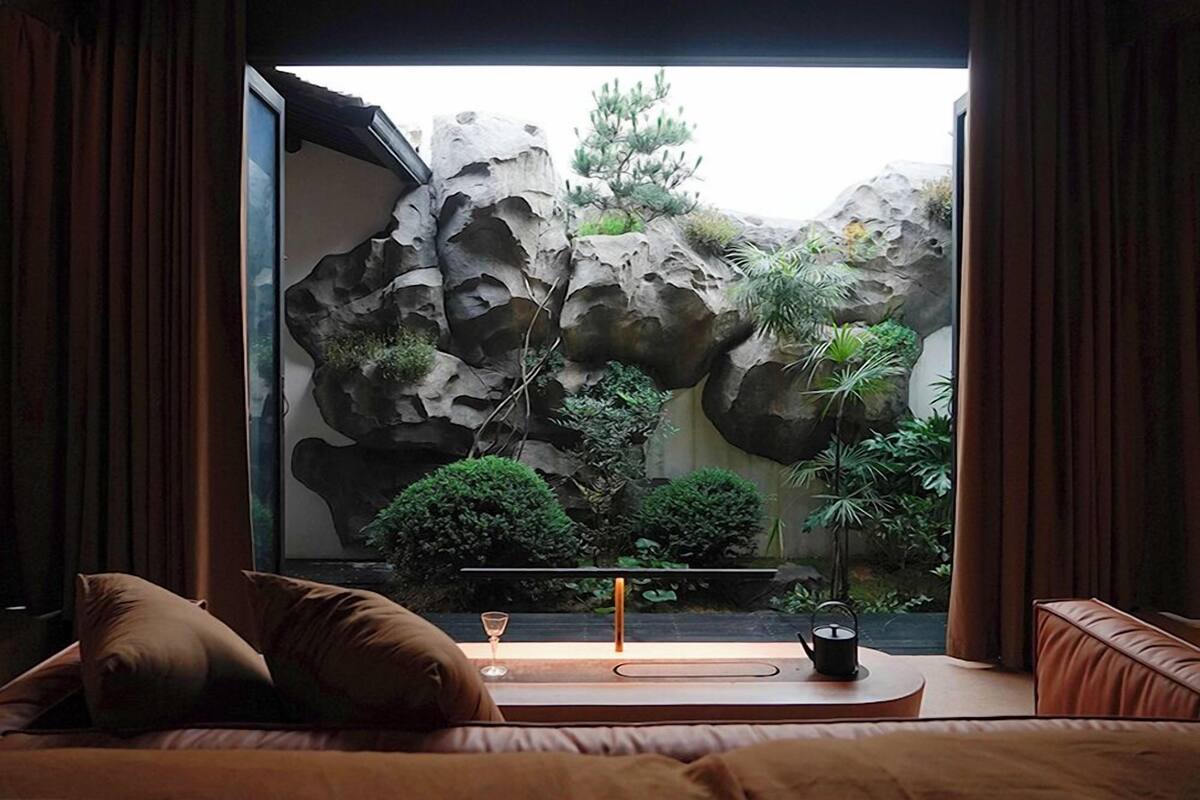 A room with a bed, a bedside table with a glass, and a view of a rock garden with plants.