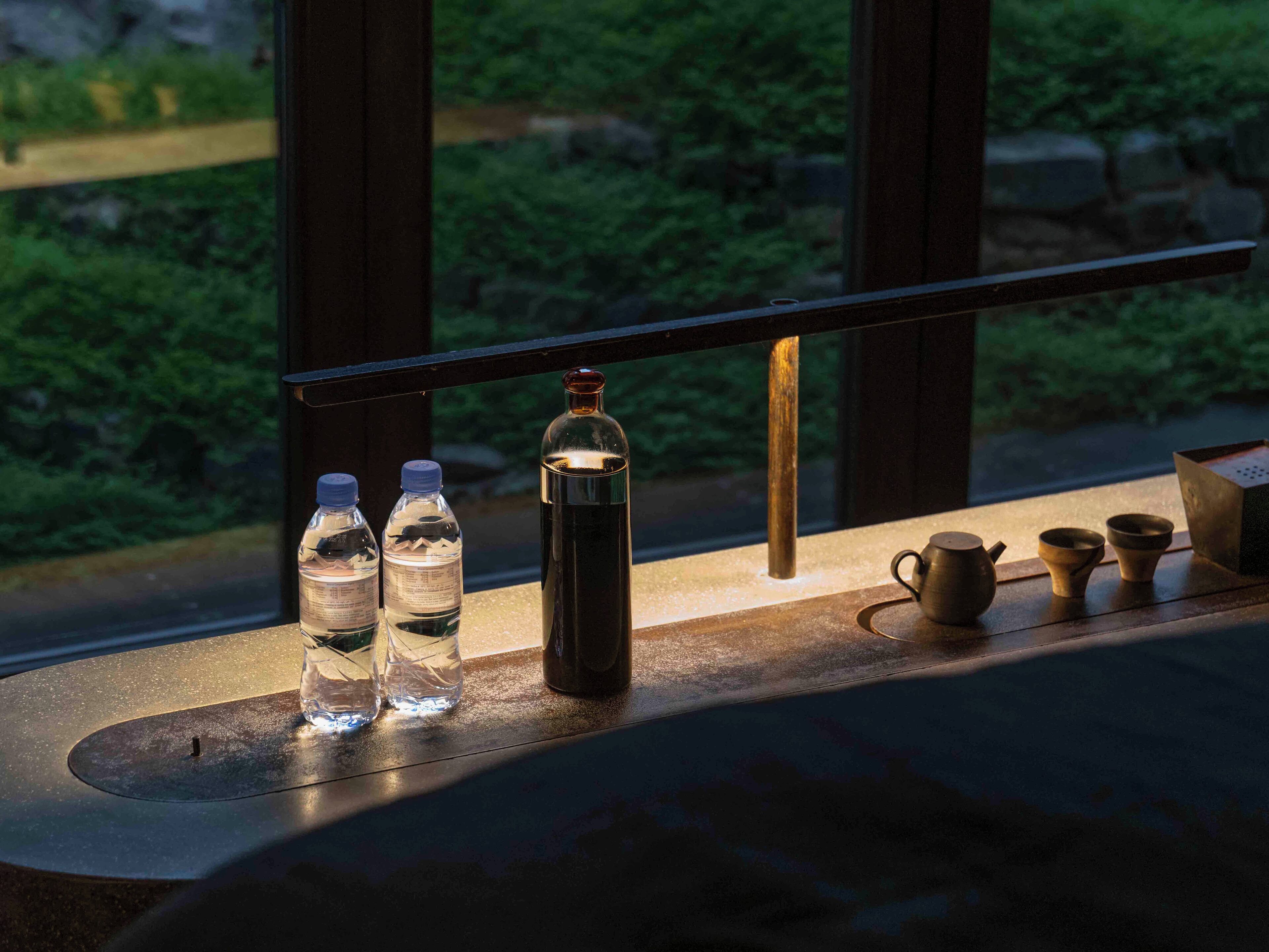 A table with water bottles, a thermos, and tea cups by a window with a view of greenery.