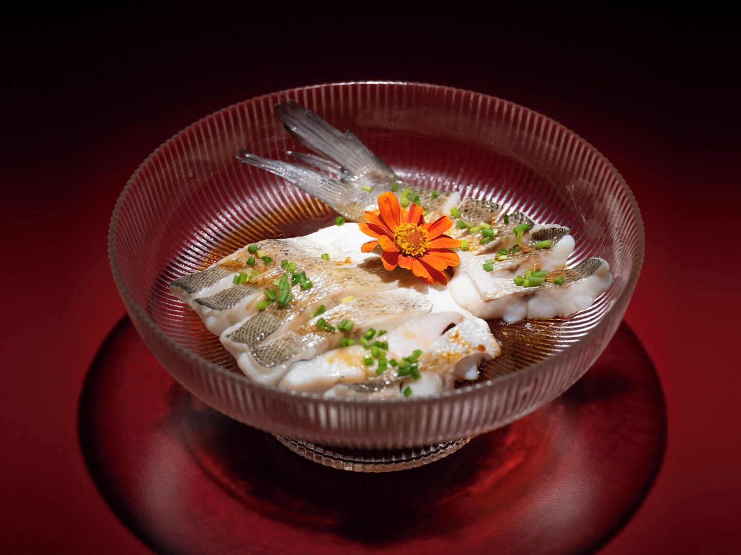 Un tazón de vidrio transparente con un plato de pescado, decorado con cebollines y una flor naranja, sobre una superficie reflectante.