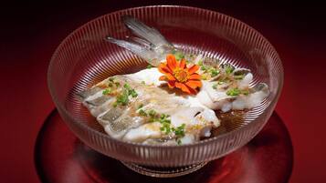 Un tazón de vidrio transparente con un plato de pescado, decorado con cebollines y una flor naranja, sobre una superficie reflectante.